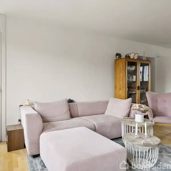 En lyserød sofa med en matchende puff står i en lys stue. Ved siden af er en træreol. Vinduer i baggrunden giver udsigt til en balkon.