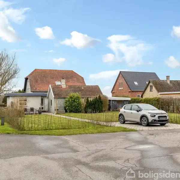 Hvid bil holder parkeret i indkørsel foran et hus med rødt tegltag; omkringliggende huse og træer under en blå himmel med skyer.