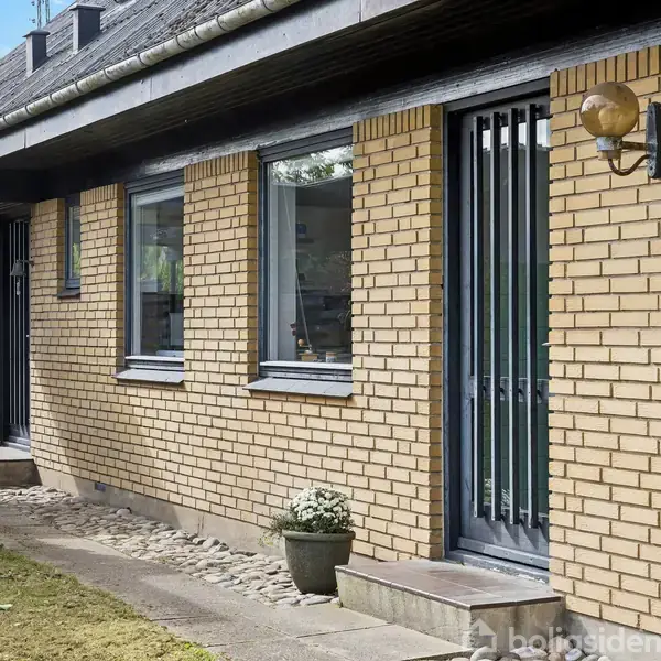 Hus med gule mursten og sort tag udsmykket med glasdøre og vinduer, omgivet af en lille have. En udendørs lampe er monteret ved siden af en dør.