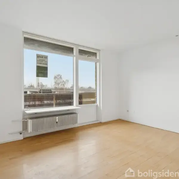 Et tomt rum med trægulv og et stort vindue, som viser udsigt til træer udenfor. En radiator er placeret under vinduet. Der er en plakat med teksten "til salg".