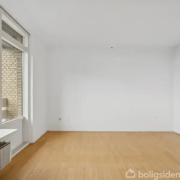 Et tomt værelse med trægulv og hvide vægge, gennemsyrer dagslys fra store vinduer til venstre. Bygningselementer ses udenfor, mens en radiator er placeret under vinduet.