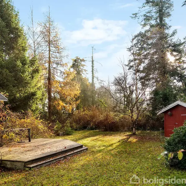En rød træbygning står stille ved en terrasse i en skovklædt have omgivet af høje træer og buske. En lille rød skur ses til højre.