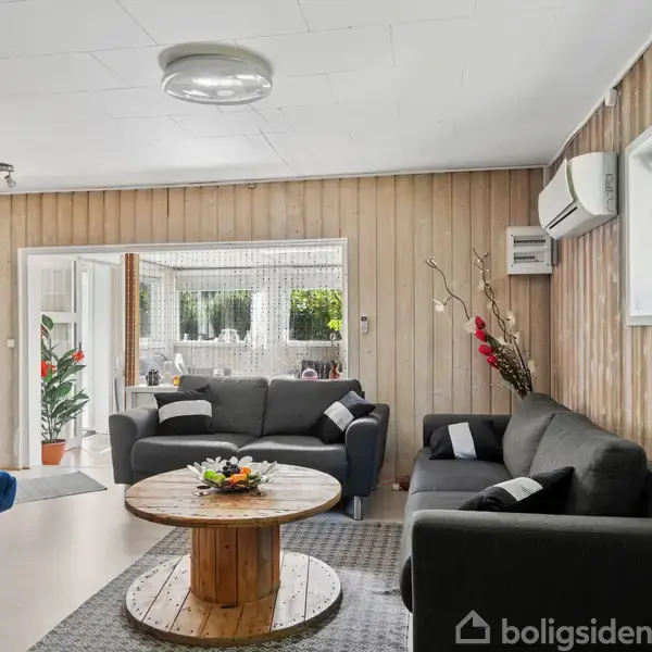 Sofagruppe med mørkegrå sofaer står i en stue, pyntet med puder. Rundt sofabord i træ midt i rummet. Væggene er træbeklædte, og et klimaanlæg er installeret.