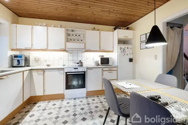 Et lyst køkken domineret af hvide skabe og en komfur i midten. Der er en spisebord med dækkede pladser og en sort lampe over. Loftet er træbeklædt.