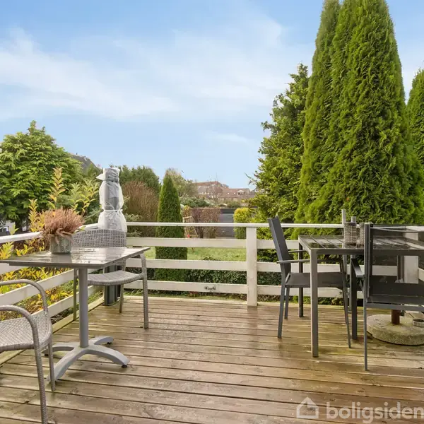 Terrasse med trægulv, hvor der står et bord med stole ved siden af en plantekrukke. Omgivet af grønne træer og buske med en klar blå himmel i baggrunden.