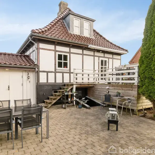 Et bindingsværkshus med mørkt tag står i en gårdsplads. Et bord med stålstole er placeret til venstre. En høj busk står ved siden af en træterrasse til højre.