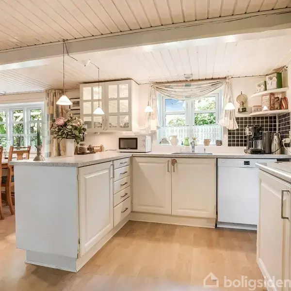 Et hvidt køkken med opvaskemaskine og mikroovn står i hyggelige omgivelser. Træbord og stole ses til venstre, med hængende lamper og vinduer, der giver naturligt lys.