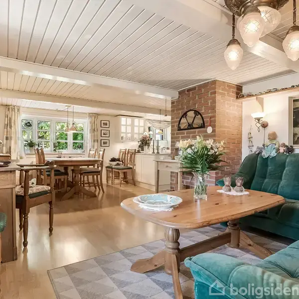 Grøn sofa står ved et sofabord med vase på tæppe; spisestue med træmøbler og lysekrone i baggrunden; lysindfald fra vinduer. Blomster og bøger dekorerer rummet.