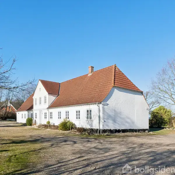 Hvidt hus med rødt tegltag står stille i en åben, solrig landskabelig område omgivet af træer og buske. En grusvej fører op til huset, og himlen er klar blå.