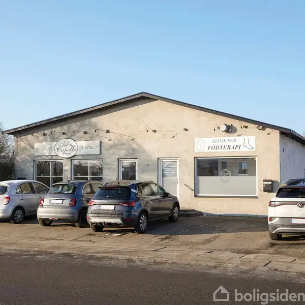 En lav, lysegrå bygning med flatskærm-tag har to skilte: "Just Beauty La" og "Klinik for Fodterapi". Fem biler holder parkeret foran bygningen på en tom vej med træer i baggrunden.