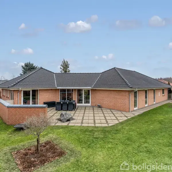 Rødt murstenshus med mørkt tag og terrasse, omkranset af en velplejet have med græs og få træer. Terrassemøbler er placeret på flisebelægningen. Skyfri himmel i baggrunden.