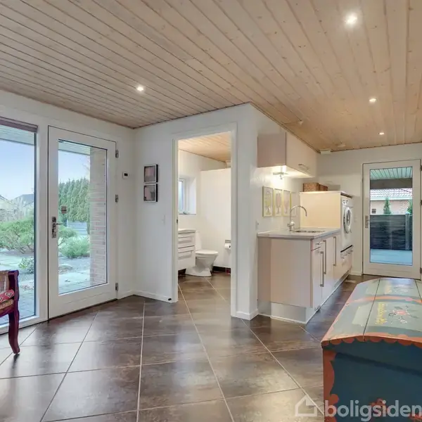 En entre med klinkegulv og træloft, stol og malerier, fører til et badeværelse og køkken med vaskemaskine. Store vinduer viser en have udenfor.