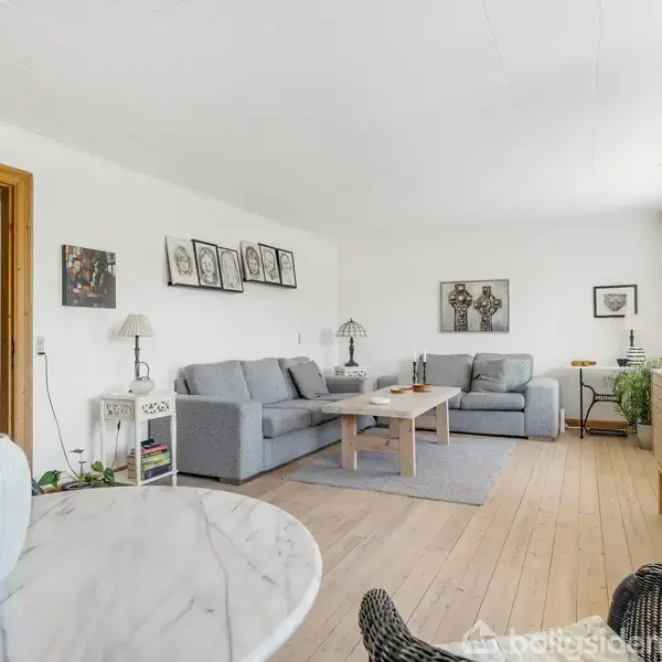Grå sofa står i afslappet position, omkranset af kunst på væggene. Stue med lyst trægulv og store vinduer lader lys skinne ind. Marbord med vase i forgrunden.