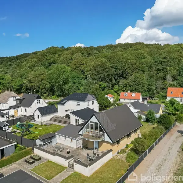 Huse med mørke tage ligger i en grøn, skovomgivet landsby. En central bygning har en rund altan og tilhørende terrasse. Små stier og haver adskiller ejendommene.