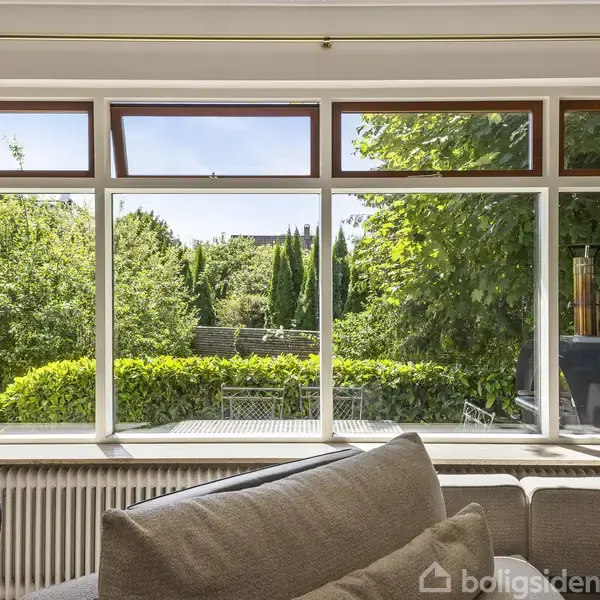 En sofa står foran store vinduer med udsigt til en frodig have. Grønt løv og hække omkranser udendørs terrassemøbler i solskin.