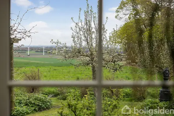 Et vinduesparti viser en have med et træ i midten, omgivet af græs og buske. I det fjerne ses en mark med vindmøller under en blå himmel.