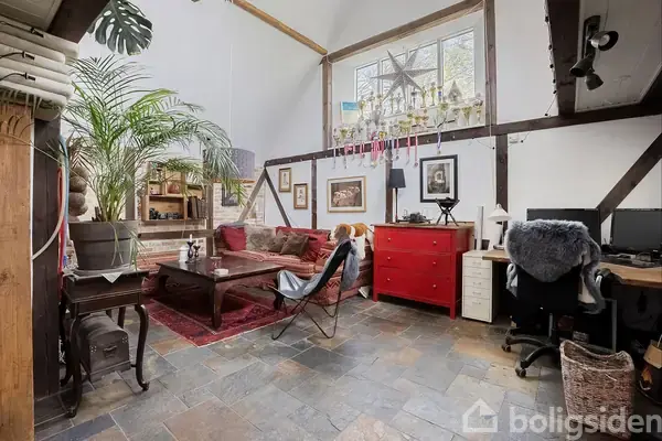 Stue med sofa og bord på rødt tæppe, omgivet af planter og kunst. Rød kommode ved vindue prydet med lysestager. Hjørne med skrivebord og stol under stor loftshøjde.