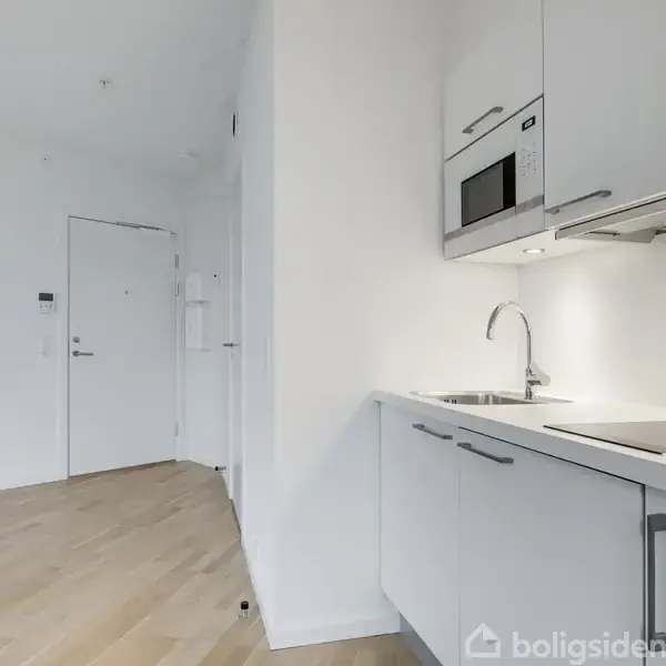 Moderne køkken med hvidt interiør, indbygget mikroovn, og keramisk komfur. Køkkenet er placeret i en lys stue med trægulv, ved siden af en hvid dør i baggrunden.