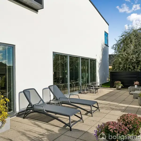 To liggestole står på en flisebelagt terrasse ved siden af en moderne hvid bygning; omgivet af potteplanter og et stakit.