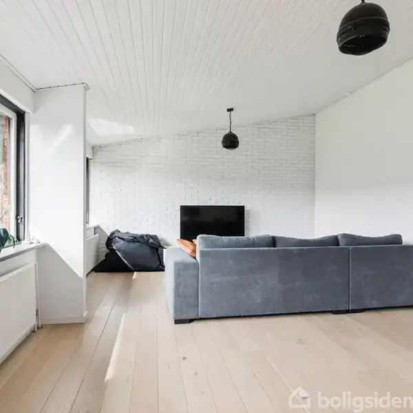 Grå sofa står foran et fjernsyn i en stue med lyse trægulve og hvidmalede vægge. Store vinduer til venstre slipper naturligt lys ind, og en sort loftlampe hænger over sofaen.