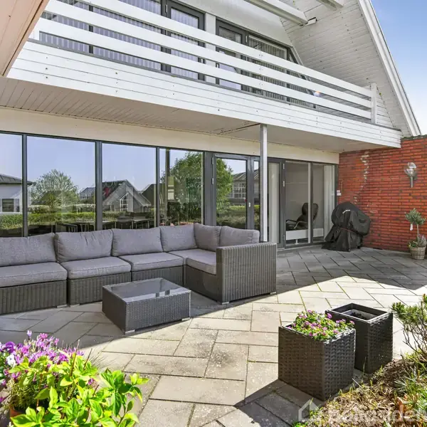 Terrasse med grå hjørnesofa og sofabord centreret, omgivet af blomsterkasser og grønne planter. Omgivelserne er et moderne hus med rødmurstenvægge og store glaspartier.