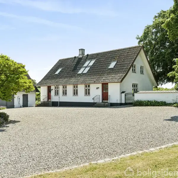 En hvid bygning med sadeltag står i baggrunden omgivet af grønne træer. Spejlingen er synlig i en stille sø i forgrunden.