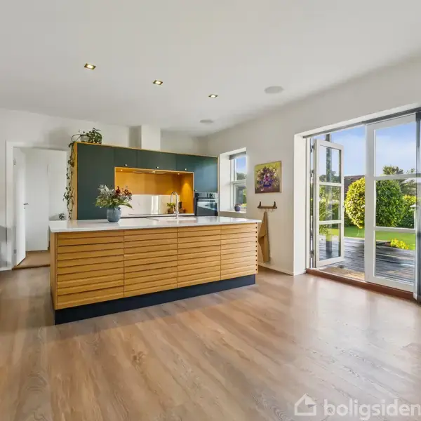 Køkkenø med træpaneler står centralt, omgivet af hvide vægge. Store glasdøre til højre åbner op til en terrasse med udsigt over en grøn have. Ingen tekst i billedet.