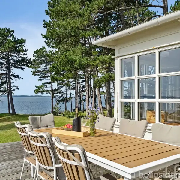 Et udendørs bord med stole står på en terrasse ved siden af et hus med store vinduer. Sammenhængende omgiver træer og udsigt til en sø eller hav.