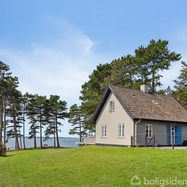 Et gråt hus med skråt tag står i en grøn have tæt på en skov af høje træer, med udsigt til vandet og en blå himmel.