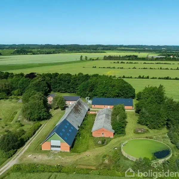 Landbrugskompleks med røde lader ligger samlet, omkranset af træer og marker. Bygningerne har blå tage, og en lille, grøncirkelformet dam er synlig ved siden af en af laderne.