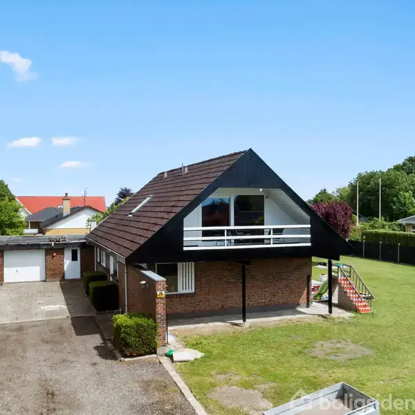 En rød murstensvilla med sort tag, balkon og en garage. Den står på en stor græsplæne omgivet af træer og en lys himmel.