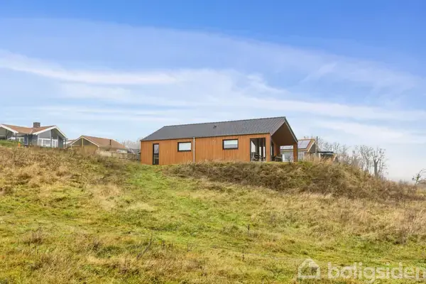 Træhus står på en grøn bakke, under en blå himmel med skyer, omgivet af græs og nogle få nabohuse i baggrunden.