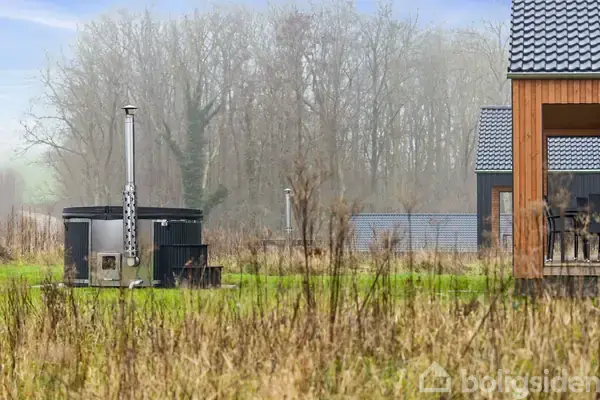 En træbygning står stille på en græsplæne omgivet af høj beplantning. En skorsten stiger op, og træer vises i baggrunden under en diset himmel.