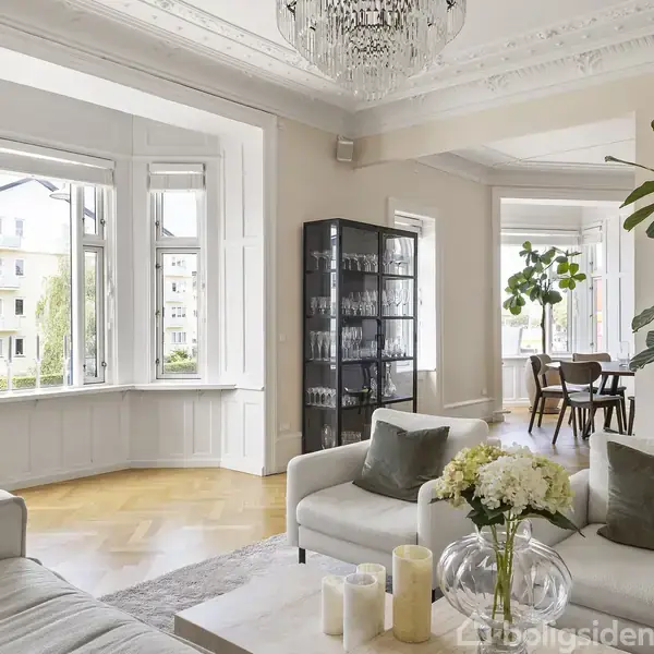 Stue med lyse sofaer og sofabord, udsmykket med planter og stearinlys. Store vinduer giver udsigt til bygninger udenfor. Spisestue med plankegulv ses i baggrunden.