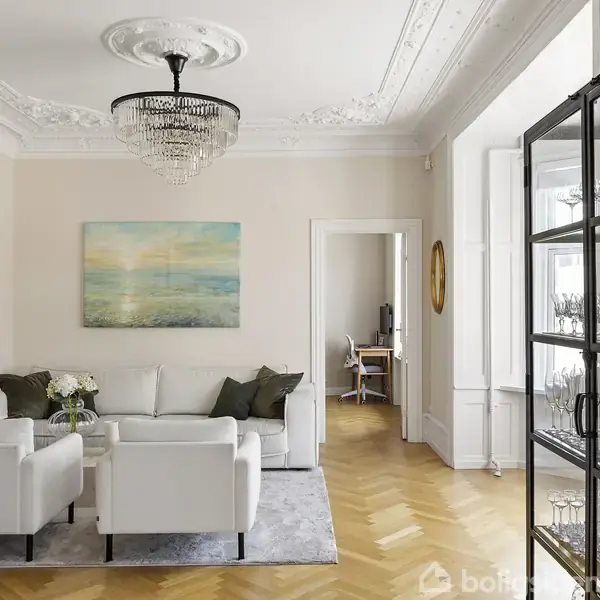 En stue med hvid sofa og stole står på et lyst trægulv. Krystal-lysekrone hænger over stuen. Maleri på væggen. Glasmontre fyldt med glas står til højre.