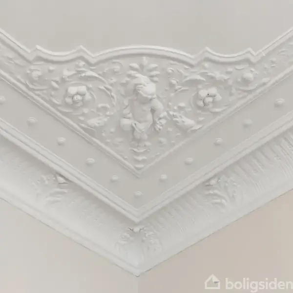 Et hvidt loftsgipsfliser viser indviklede, dekorative mønstre med blomster og blade. Det er en udsmykning, der løber langs loftets hjørne mod en beige væg.