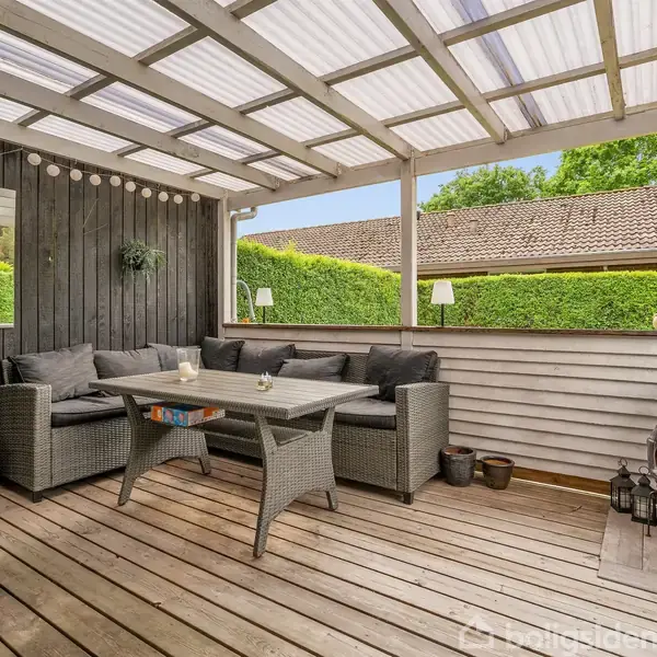 Terrassemøbler står under et halvtag, bestående af en grå sofa og et bord. Baggrunden har grøn beplantning og et tag i træ omgiver området. Lanterner er placeret rundt omkring.