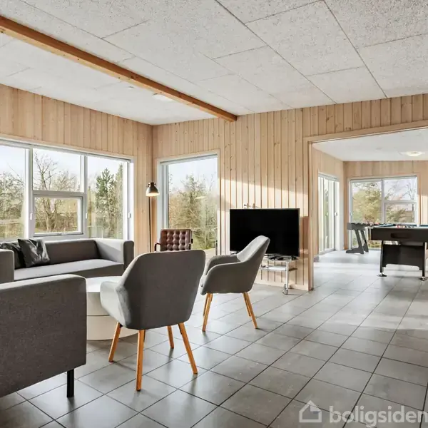 Stue med moderne sofaer og lænestole placeret på gråt flisegulv, set mod store vinduer og en fladskærms-TV. Forestil dig et lyst rummeligt rum med træpanelvægge.