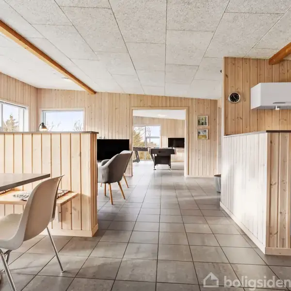 Et åbent rum med lyse trævægge og gråt flisegulv. En bordgruppe til venstre, køkken i højre hjørne, og en hyggelig stue længere fremme med store vinduer.