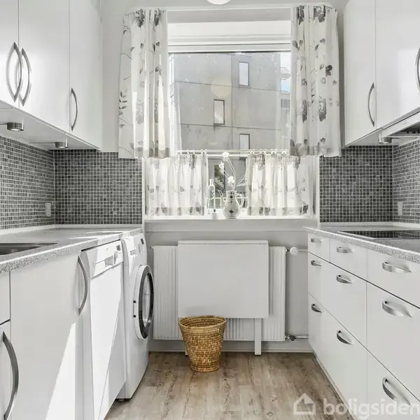 Et lyst køkken med hvide skabe og grå flisemønster på væggene. En vaskemaskine og ovn er indbygget. Et vindue med blomstrede gardiner kigger ud til en grå bygning.