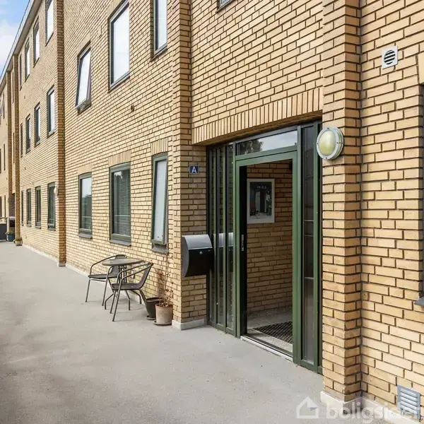 En murstensbygning med åbne døre og et tilsluttet gangareal. To udendørs stole og planter er ved siden af indgangen. Træer ses i baggrunden.