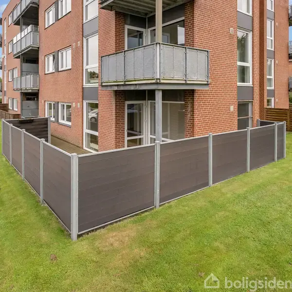 En rød murstensbygning med altaner omgivet af en hæk. Foran bygningen er en indhegnet terrasse. Græsplæne omgiver området, der er i et beboelsesområde.