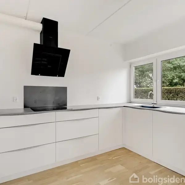 Et moderne, hvidt køkken med komfur og ovn hængende på væggen. Store vinduer giver udsigt til en grøn have. Gulvet er lyst træ.