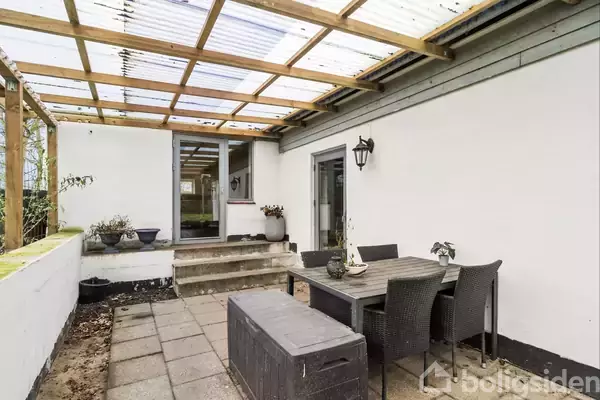 En overdækket terrasse viser et havebord med stole, omgivet af hvide vægge og krukker med planter. En lysegrå boks står ved siden af bordet.