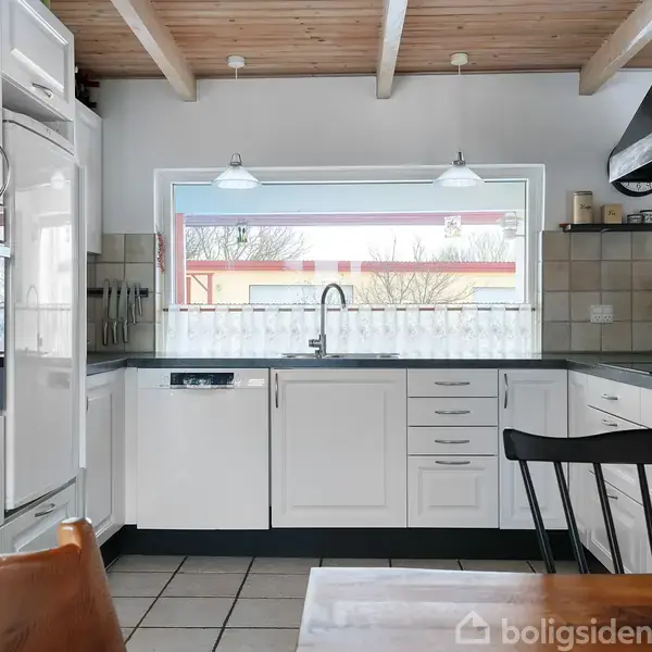 Hvidt køkken med ovn og opvaskemaskine. Der står en sort emhætte over kogepladen. Træloftet har synlige bjælker. Store vinduer giver udsigt til et hus udenfor.