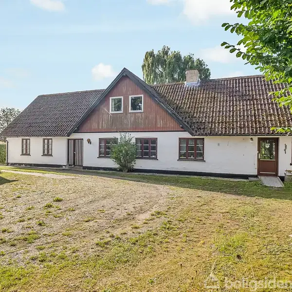 Et stort, hvidt landhus med brun tag er omkranset af en græsplæne og træer på en solskinsdag. Til venstre ses en mindre udbygning med skråt tag.
