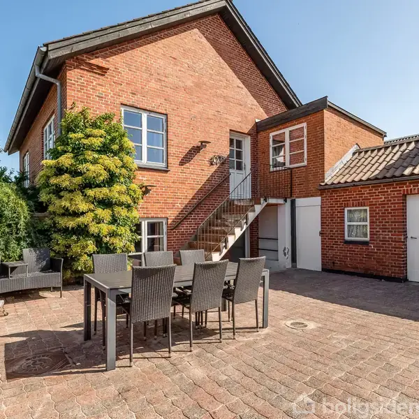 Rødt murstenshus med en udendørs terrasse, hvor et spisebord og stole står. En trappe leder til en dør på første sal. Omgivet af høj vegetation under blå himmel.