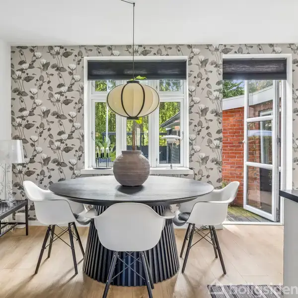 Rundt spisebord med seks hvide stole, placeret i et lyst rum med blomstrede tapetvægge og store vinduer. En stor vase står midt på bordet under en hængende lampe.