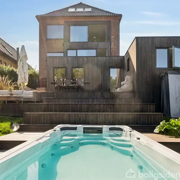 Et moderne hus med store vinduer står bag en pool i en velplejet have. Der er en terrasse med sofaer og parasoller.