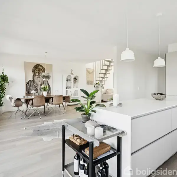 Et moderne, lyst køkkenalrum med en hvid køkkenø i forgrunden. Bagved ses et spisebord med stole og en kunstplakat. Rummet har store vinduer og planter.